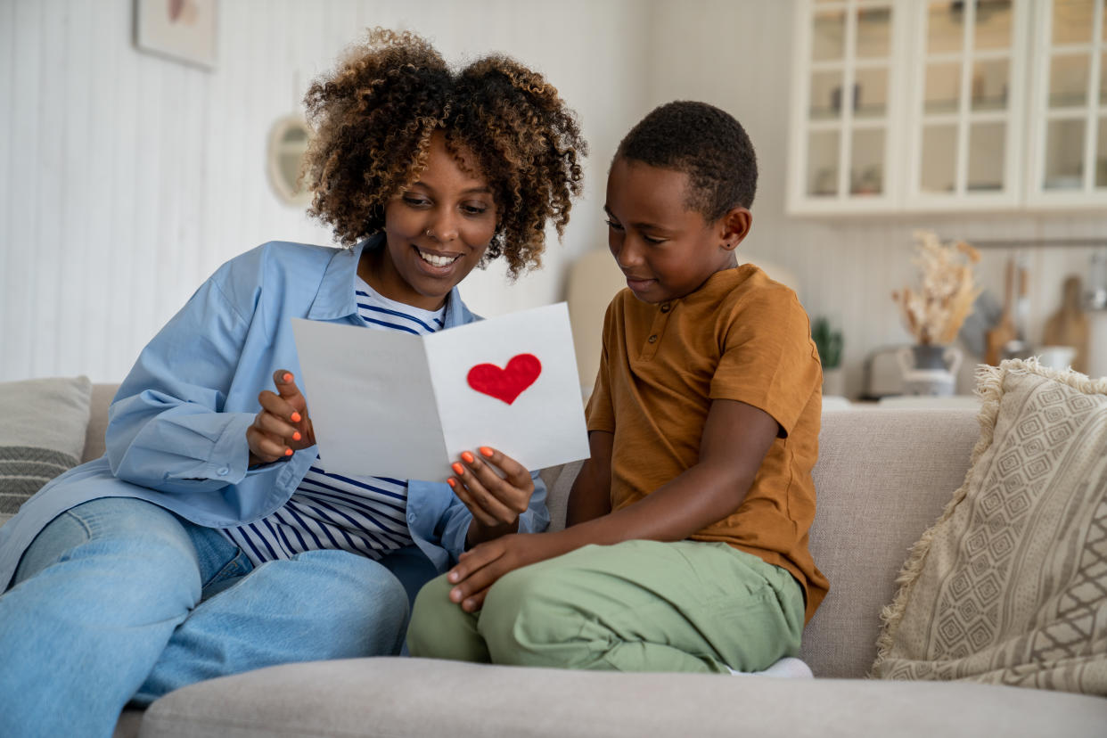 A woman and her young son sit on a couch together and look at a Valentine's Day card with a red heart on the front.