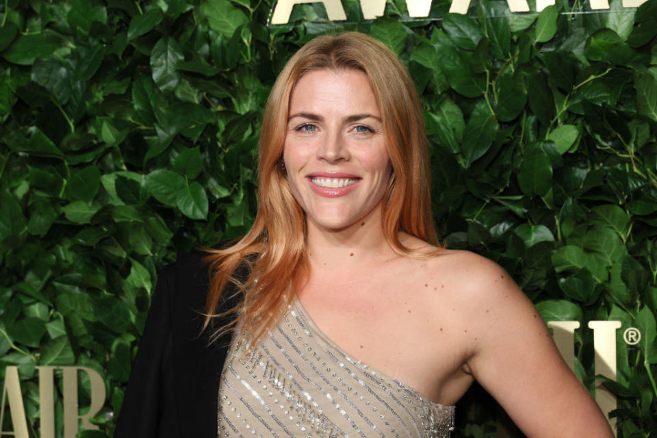 A close-up of Busy smiling and wearing a one-shoulder outfit against a leafy background