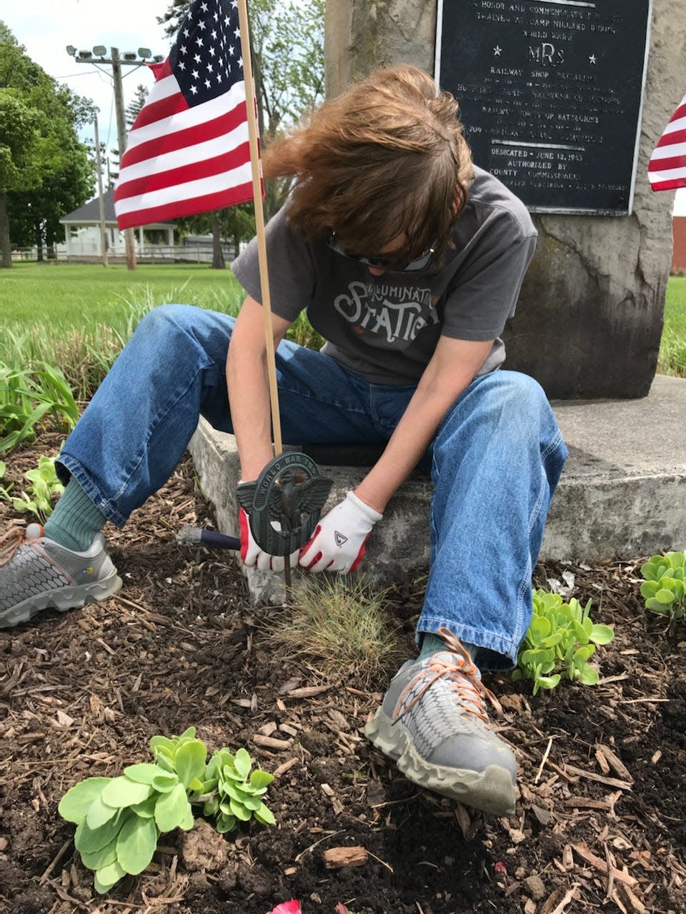 The Camp Millard Memorial marker is one of the "Company's Coming" landscaping efforts at the Crawford County Fairgrounds. In an older photo, Janet Nance scrapes paint from the concrete base. The Earth, Wind and Flowers Garden Club maintains this plot.