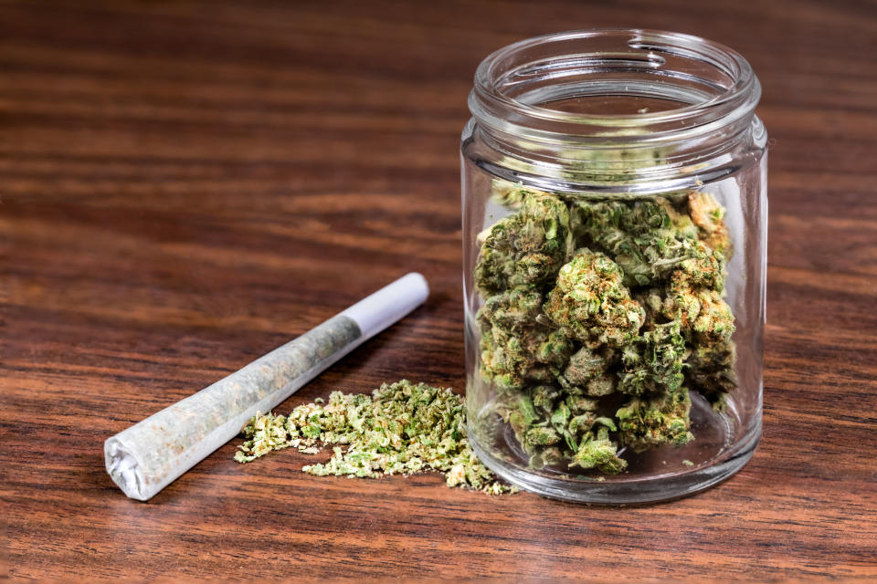 Dried cannabis flowers in a jar sitting on a wooden table next to a rolled combustible