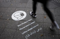 A pedestrian walks over a reminder to wear face masks sprayed on the sidewalk at the Karl-Marx-Strasse at the district Neukoelln in Berlin, Germany, Tuesday, Oct. 27, 2020. Karl-Marx-Strasse is one of the streets in the German capital where face masks are mandatory to prevent the spread of the coronavirus. (AP Photo/Markus Schreiber)