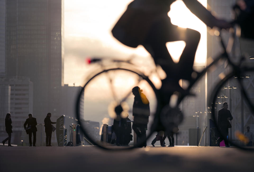 People walking and cycling in an urban environment at sunset, with a city skyline in the background. The silhouette of a cyclist in the foreground