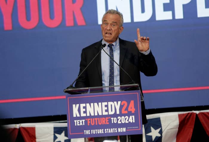 RFK Jr. at podium with "Kennedy 24" sign, gesturing with one hand while speaking