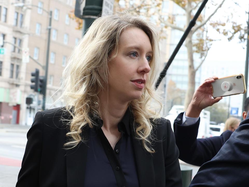 Elizabeth Holmes wearing a black jacket while standing next to a group of reporters on a city street.