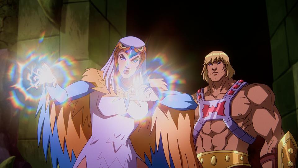 In a CG animated still from Masters of the Universe: Revelation, He-Man (right) stands behind Sorceress (left) in a stone room lit only by the light coming from SorceressesÕ hands. Sorceress has her palms outstretched in front of her and wears a white dress, a falcon-like hood over head and orange and blue wings. He-Man wears a silver chest plate with a red "H" in the center, a golden armored belt and wrist plates.