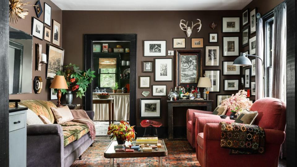 gallery wall of pen and ink drawings, with antlers hung above, in living room painted dark brown with rust red club chairs, gray couch, turkish style rug in autumnal tones, in 1940 craftsman cottage owned by leighton, alabama, landscape gardener anthony brewington