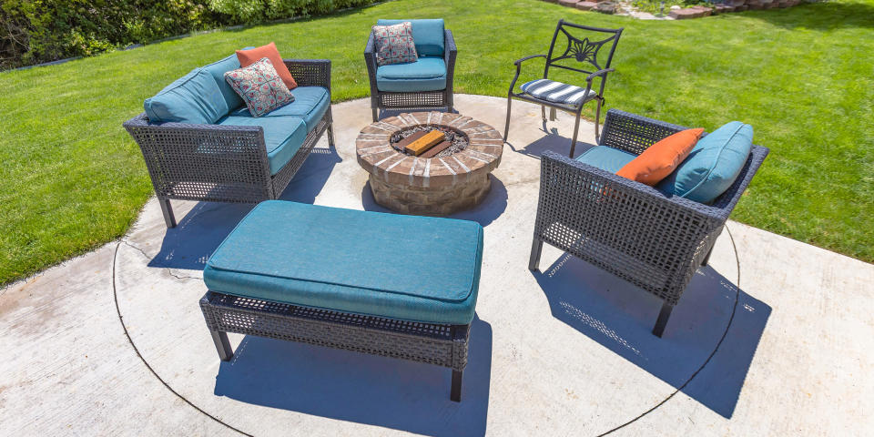 Circular fire pit and chairs on a sunny backyard. A circular stone fire pit surrounded by chairs with blue cushions and pillows. The fire pit is on a backyard patio with landscaped ground.