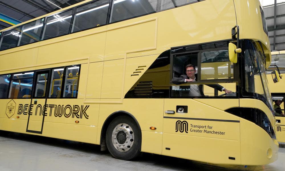 Artistically angled photo of a sleek modern double decker  bus in canary yellow, with Andy Burnham leaning out of the driver’s cab window