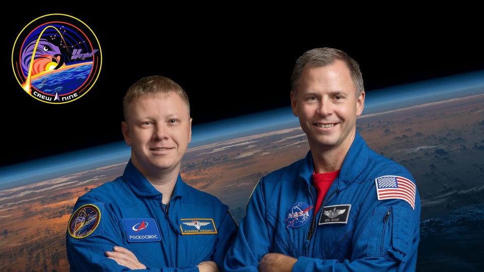   Dos hombres con trajes de vuelo azules posan para un retrato con los brazos cruzados frente a un fondo de la Tierra vista desde el espacio. 