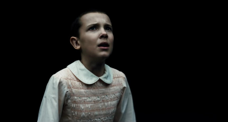 Eleven from Stranger Things appears worried in a light-colored dress with a ruffled collar