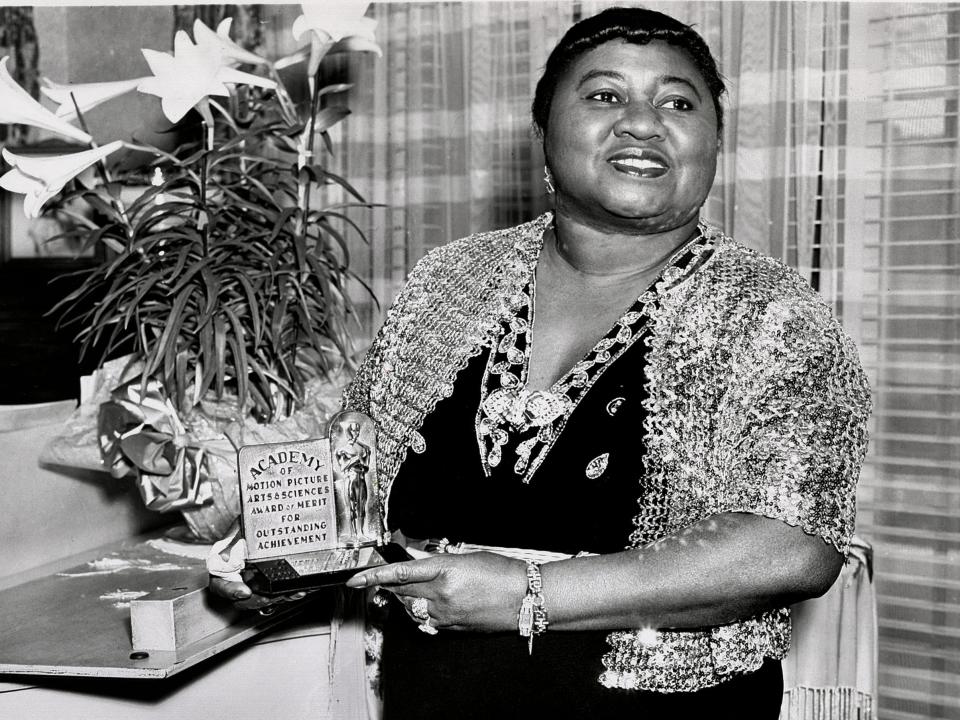 Black and white photo of Hattie McDaniel in a dress smiling and holding her award plaque.