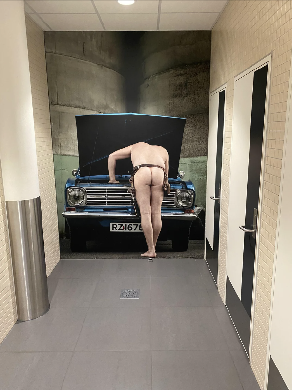 Mural of a person wearing minimal attire bending over, inspecting a car engine. Positioned in a hallway between bathroom doors
