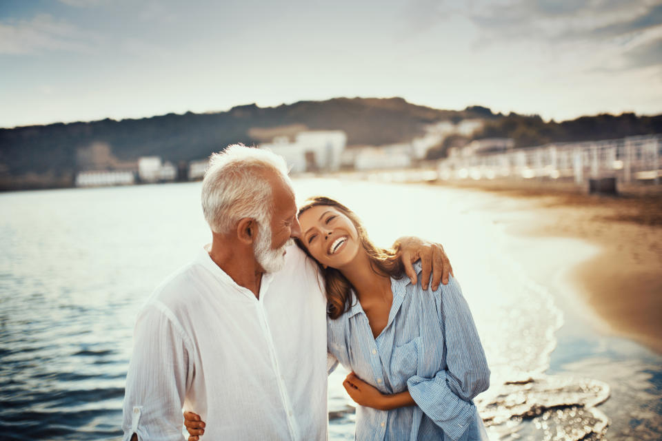 An elderly man and a young woman, both smiling and wearing casual shirts, walk arm-in-arm along a beach with their heads close together