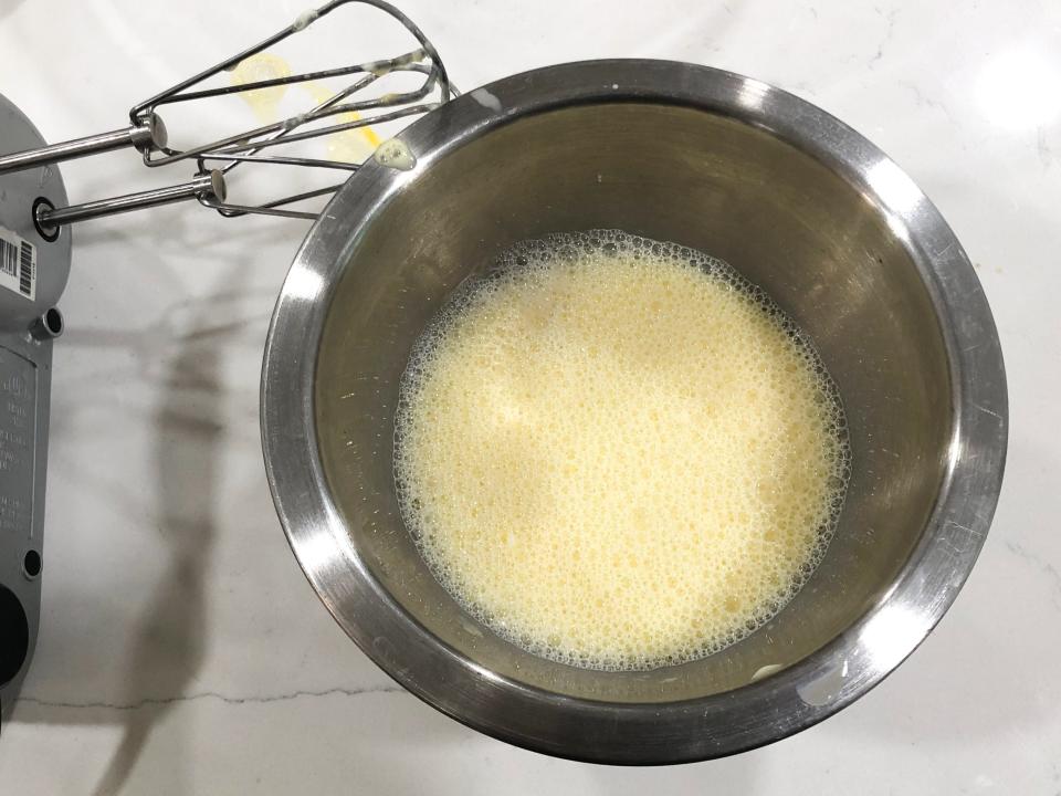 An electric hand mixer next to a silver bowl with yellow liquid. The mixer and bowl are on a white counter.