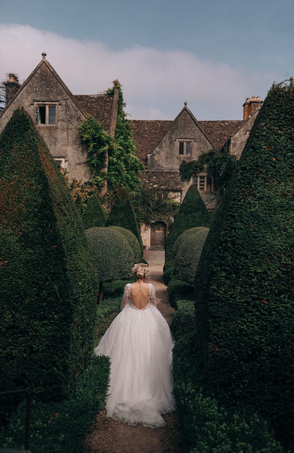 A person in a wedding dress walks through a garden in front of a mansion.