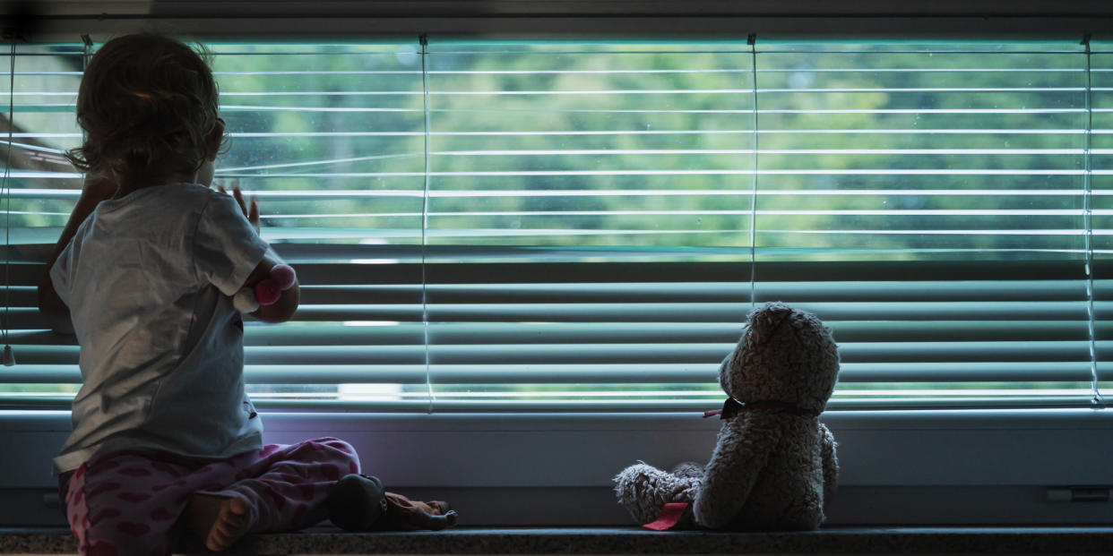 Toddler girl looking out the window with her bear beside her. 