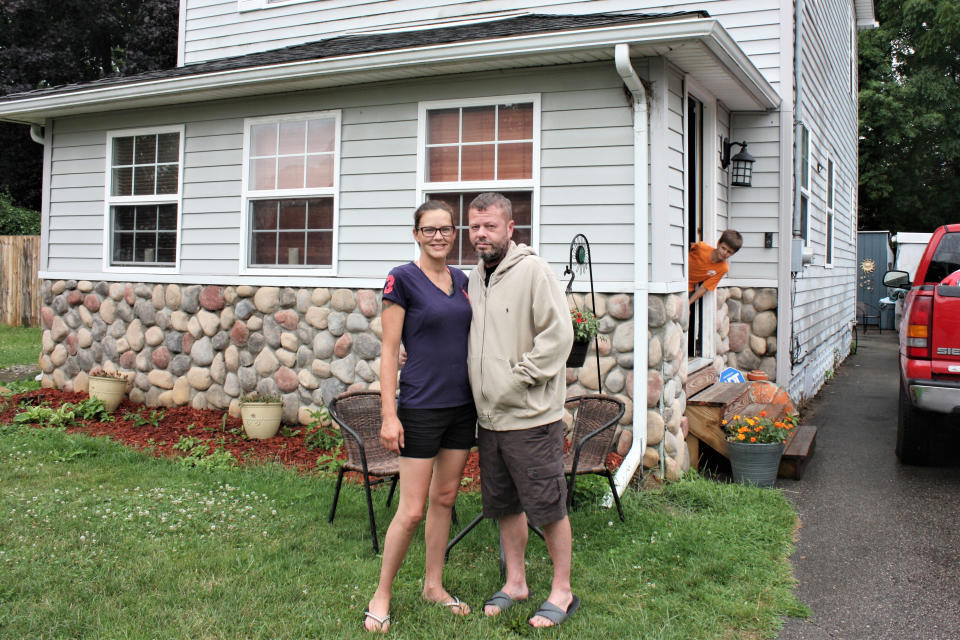 Crystal and Chris Martin stand outside their home, Sunday, July 19, 2020 in Burton, Mich., as one of their children looks on. The Martins, who had to defer some mortgage payments, are among millions of Americans who have struggled financially during the coronavirus pandemic. Crystal has been laid off since March from her job at a roller skating rink and Chris, an X-ray technician at a Flint hospital, was laid off, then took parental leave after the birth of a child this month. A new study by NORC at the University of Chicago finds that more than a quarter of Americans report losing income, investments and savings during the pandemic. (AP Photo/Tammy Webber)