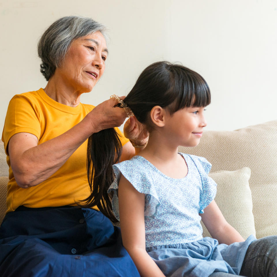 An older woman is braiding the hair of a smiling young girl who is sitting on a couch. Both appear happy and content