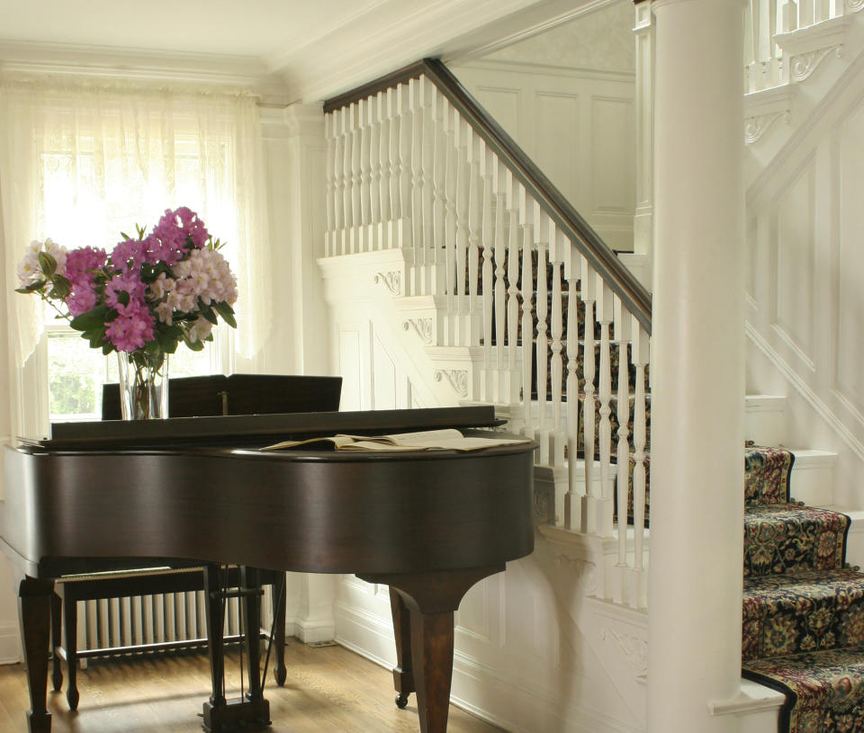 A grand piano with sheet music sits next to a staircase adorned with a floral-patterned carpet. A vase of flowers is on top of the piano. Natural light filters through curtains