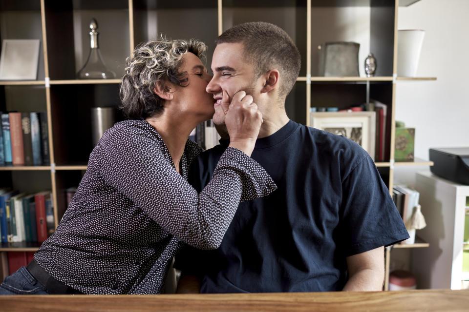 Person with short, curly hair kisses a smiling man on the cheek, squeezing his face gently