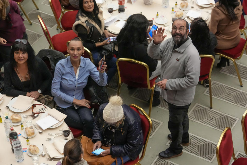 Don Andrea Conocchia, right, waves a s he stands next to a table with a group of transgender women he accompanied at a lunch for the poor with Pope Francis, in the Paul VI Hall at the Vatican, Sunday, Nov. 19, 2023. Pope Francis offered several hundred poor people, homeless, migrants, unemployed a lunch on Sunday as he celebrates the World Day of the Poor with a concrete gesture of charity in the spirit of his namesake, St. Francis of Assisi. (AP Photo/Andrew Medichini)