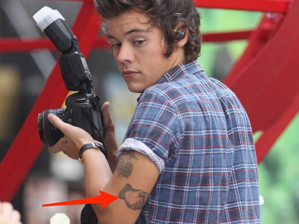 A red arrow pointing to a tattoo of two hands shaking on Harry Styles' arm.