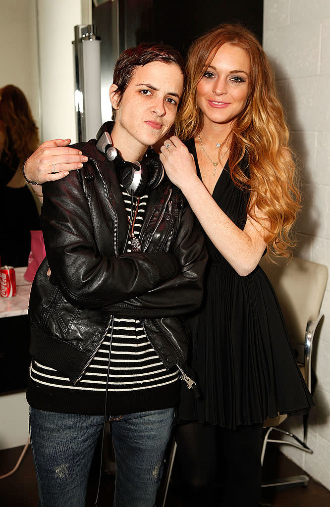 Samantha Ronson in a leather jacket with headphones around her neck, and Lindsay Lohan in a dress pose together, smiling gently with Lohan's hand on Ronson's shoulder