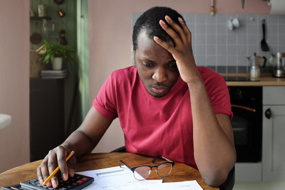 Man sitting at table, holding head while looking at documents and typing on calculator