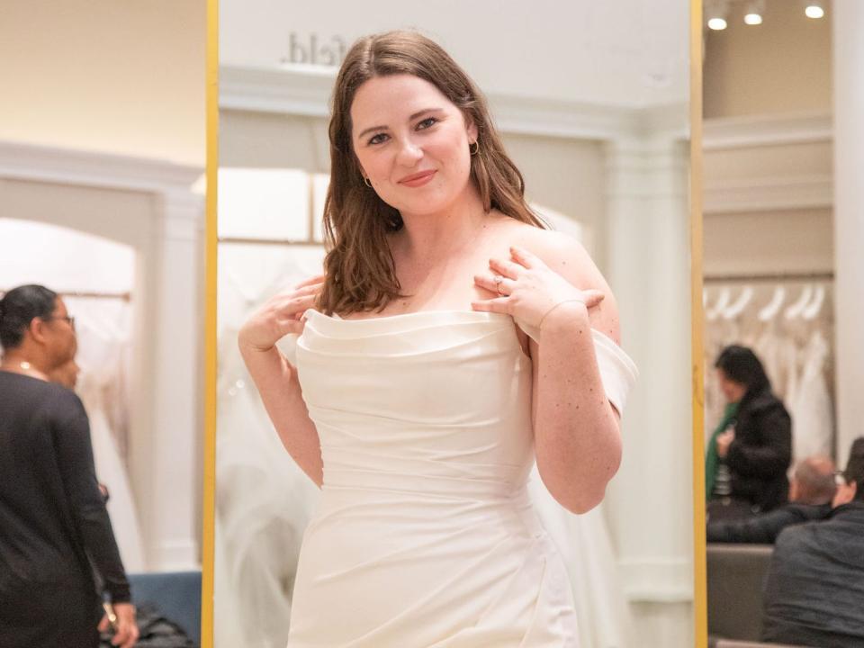 A woman stands in a wedding dress with a mirror behind her.