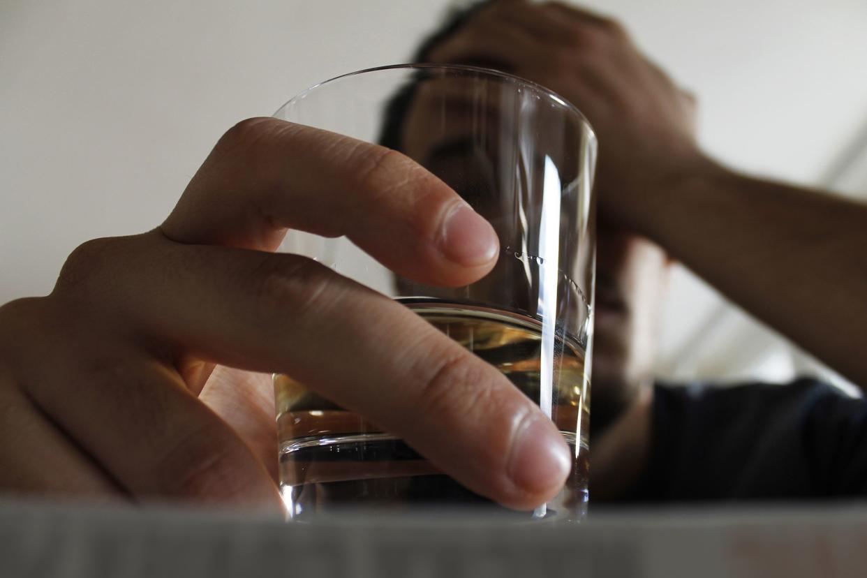 Closeup of fingers holding shot glass with whiskey on a table in the foreground, with a person holding his head with the other hand, blurred in the background, a light grey wall in the background