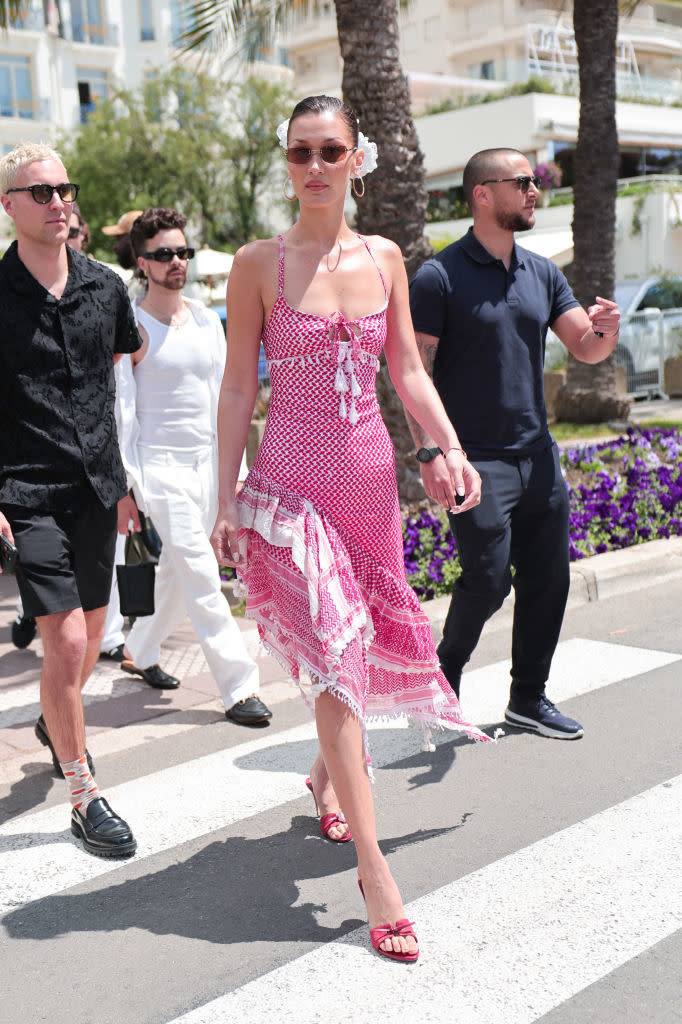 Bella Hadid walking outdoors with three companions, wearing a red and white sundress, sunglasses, and red sandals