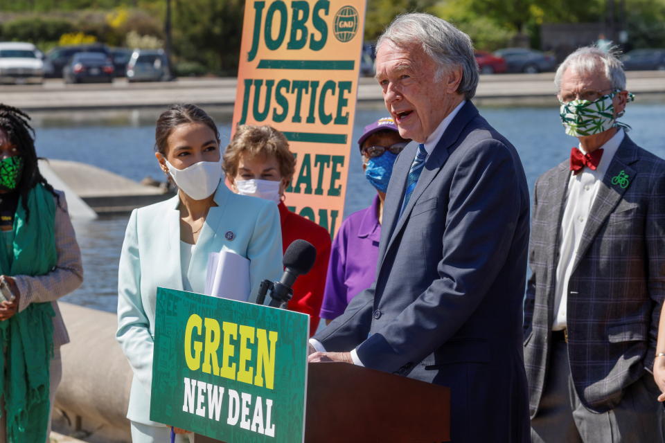 Representative Alexandria Ocasio-Cortez watches from the side as Senator Ed Markey speaks at a podium at a news conference.