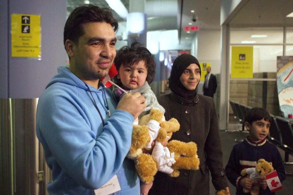 A man holds a baby and two teddy bears and smiles as his smiling wife and son, holding a small Canadian flag, stand beside him.