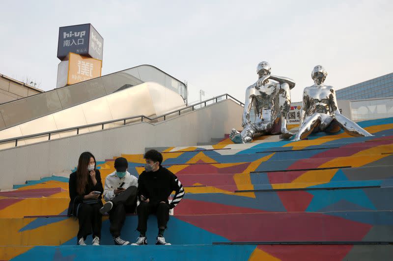 People wearing face masks sit on steps in front of a public sculpture at a shopping complex in Beijing