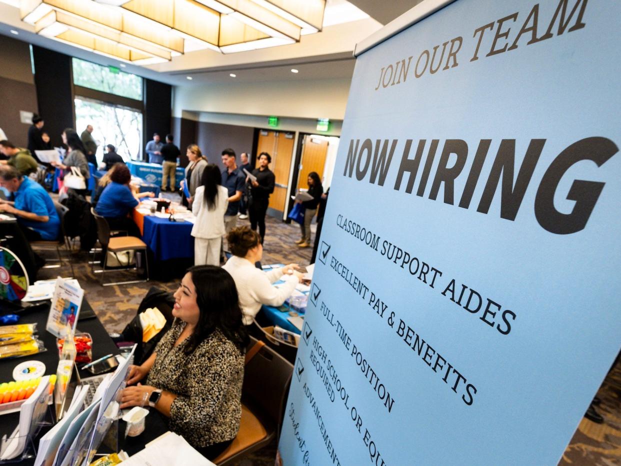 A sign that says now hiring and people at a career fair