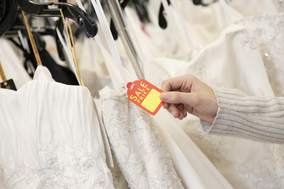 Hand holding a "Sale Price" tag on a wedding dress among several dresses on hangers