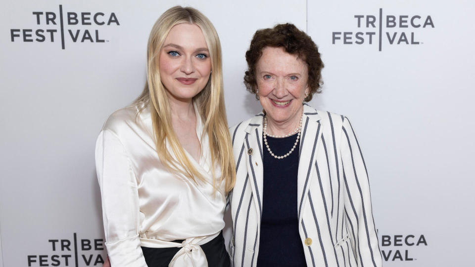 Actor and producer Dakota Fanning wearing a white blouse and standing with an older woman in a black shirt and white pinstriped blazer in front of a white backdrop for the Tribeca Festival. 