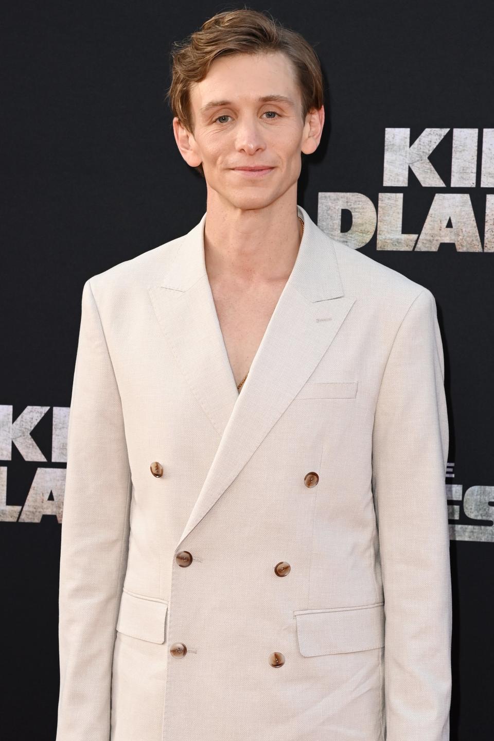 Travis Jeffery in a double-breasted cream suit stands smiling at a premiere event for the Kingdom of the Planet of the Apes