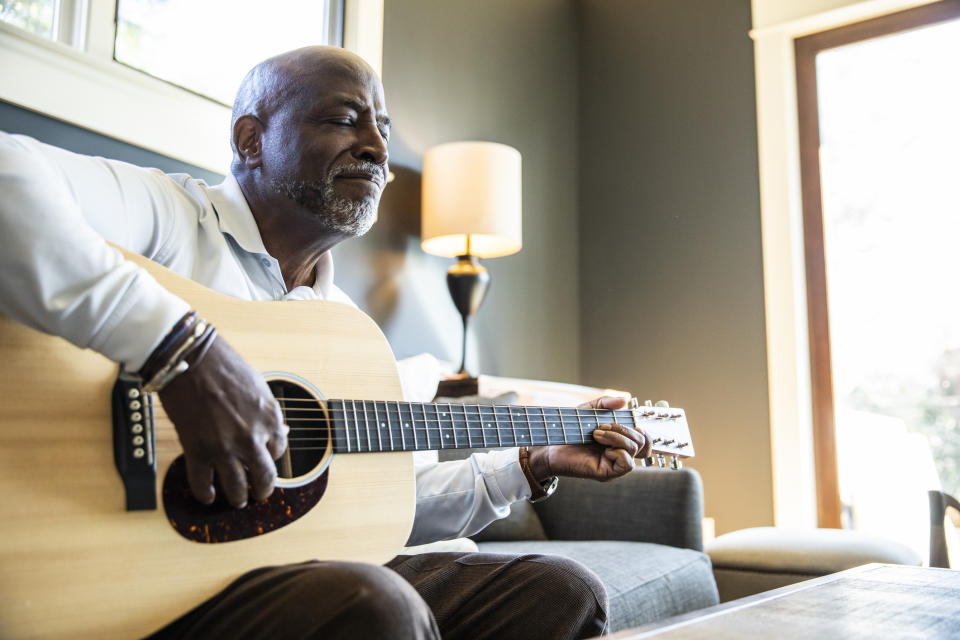 Man smiling while playing acoustic guitar, seated in a well-lit room