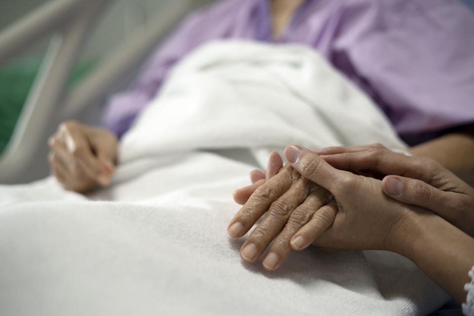 An older adult patient lying in a bed holding the hand of a loved one in the hospital. (Photo via Getty Images)