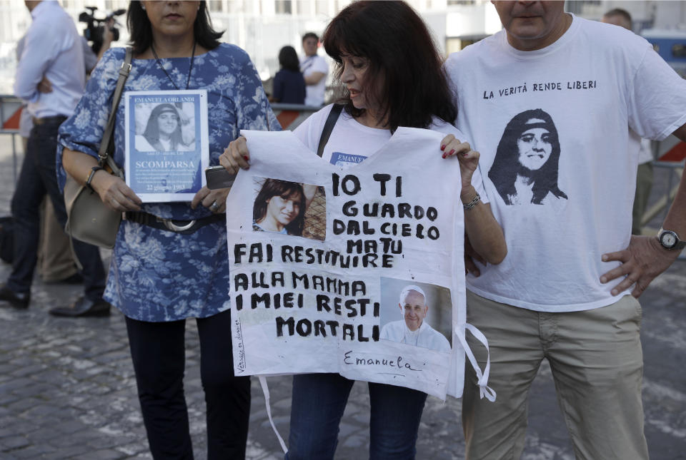 Reanta Grattani, who claims to be a friend of the Orlandi family, center, holds a t-shirt with the pictures of Emanuela Orlandi and Pope Francis and a writing reading: I'm watching you from the sky, but you (referring to Pope Francis) please let them return my mortal remains to my mother, as she gathers with Cinzia Di Florio, left, holding a picture of Emanuela reading: Missing, and Sandro Masetti wearing a ti-shirt with a picture of Emanuela reading: The Truth will set you free, outside the Vatican, Saturday, July 20, 2019. The mystery of the 1983 disappearance of the 15-year-old Emanuela Orlando, daughter of a Vatican employee, took yet another twist Saturday as the Vatican formally opened two ossuaries discovered under a stone slab. The boxes of bones were found after the Vatican opened the tombs of two 19th-century German princesses in the cemetery of the Pontifical Teutonic College in hopes of finding the remains of Emanuela Orlandi. (AP Photo/Gregorio Borgia)