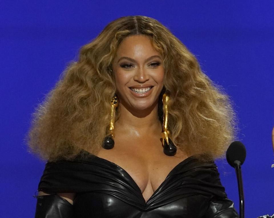 A smiling woman with long curly hair and dangling earrings stands at a microphone.