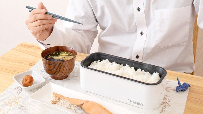 A person sitting at a desk eating from an electric bento box full of rice using chopsticks.