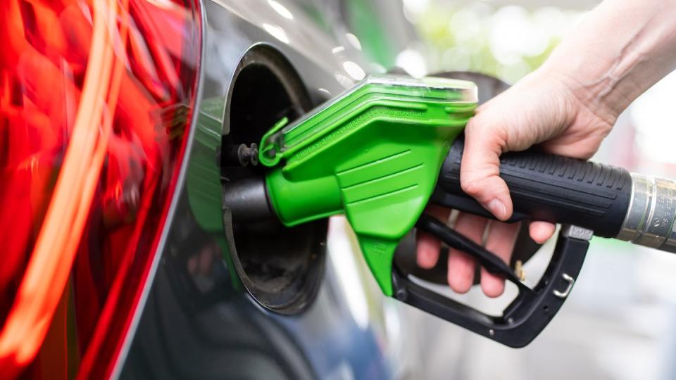 Eine Frau hält an einer Tankstelle an einer Zapfsäule eine Zapfpistole in der Hand und betankt ein Auto.