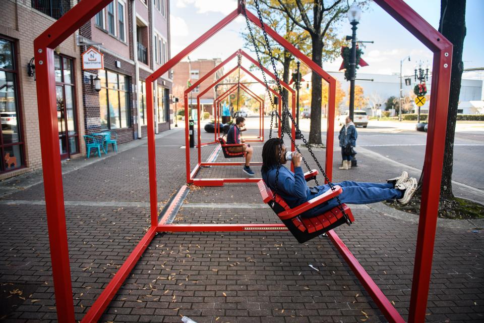 Tre Webb takes a sip of coffee while swinging on the public-interactive art exhibit, called Mi Casa, Your Casa 2.0, on Hay Street, Monday, Nov. 28, 2022. The art exhibit is made up of eight art pieces, which are located on Hay and Person streets, showcases red swings shaped as small houses. When the houses are empty, the glow softly white, but when a visitor steps inside, the white glow intensifies.