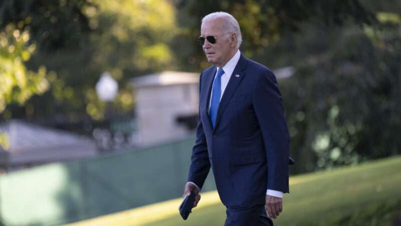 President Joe Biden wearing a suit and sunglasses walks past a background of green grass and green trees.