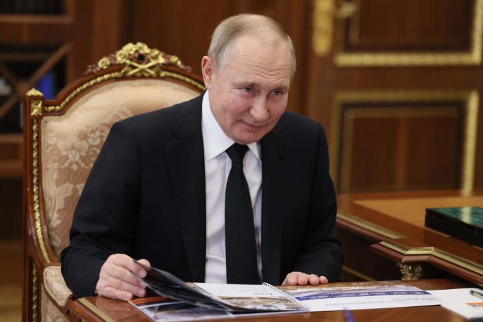 Vladimir Putin seated in an ornate chair at a desk in a dark suit and tie with hands on a thin spiral-bound booklet.