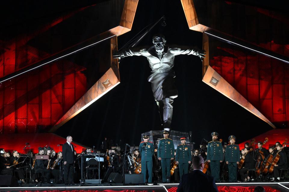 Vladimir Putin walks across a stage with soldiers and an orchestra, with a large statue in the background.