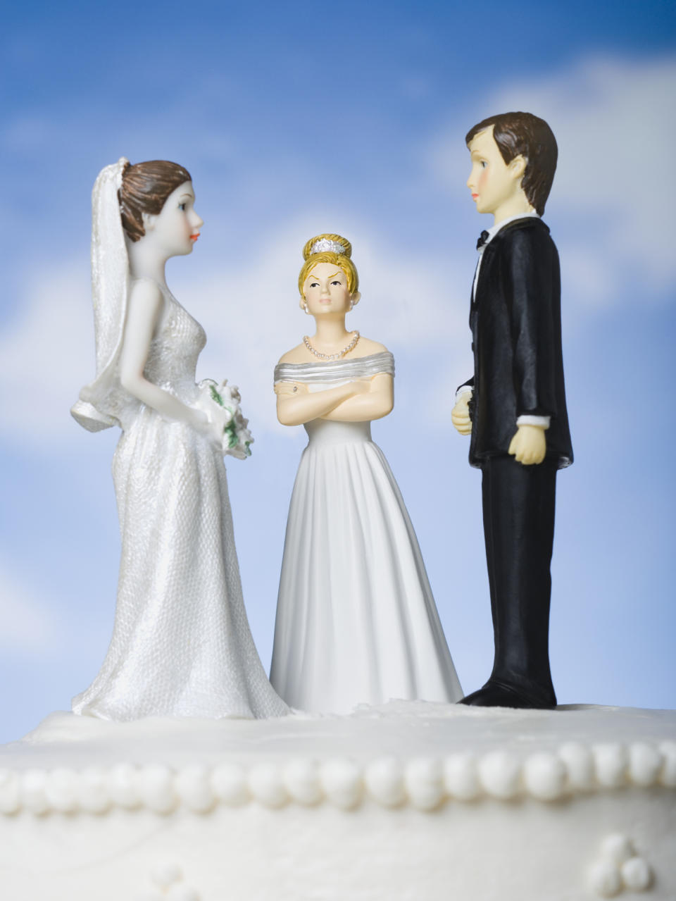 Wedding cake toppers depicting a bride and groom facing each other with an angry woman in a white dress and tiara standing between them, her arms crossed