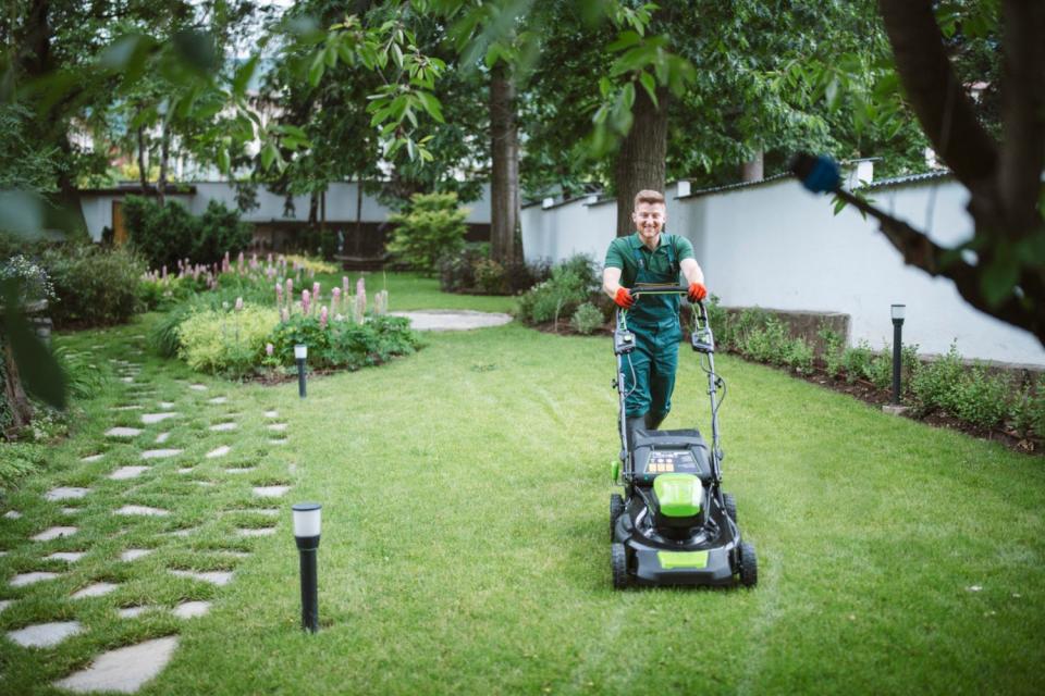 A worker in a green uniform pushes a lawn mower across a green lawn. 